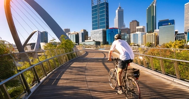 Cyclist riding on a boardwalk near the Elizabeth Quay bridge with Perth's skyline in the background.