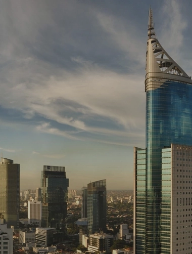 Panoramic city skyline with a tall, curved blue-glass skyscraper at center, surrounded by high-rise buildings, streets, and dense urban neighborhoods under a partly cloudy sky at golden hour