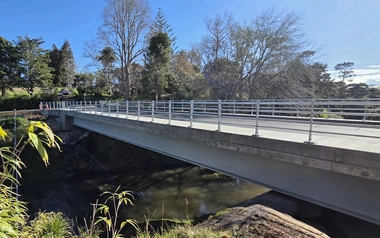 Concrete bridge spanning a stream with railings and trees along the riverbank.