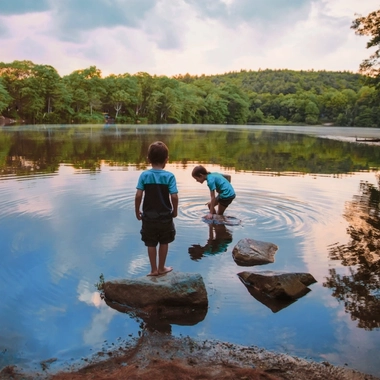 Two children standing and playing in the shallow water of a calm lakeshore surrounded by trees at sunset