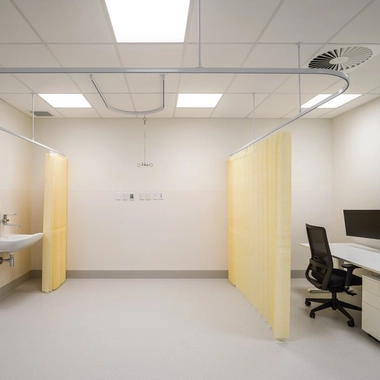 Medical consulting room with reclining chair, hand‑washing station, yellow privacy curtains, and dual computer workstation.