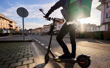 Person with a delivery backpack riding an electric scooter on a sunlit suburban street.