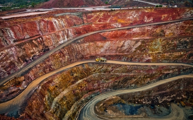 Aerial view of an open-pit mine with winding roads and a mining truck navigating steep paths. Vibrant earth layers show rich mineral deposits.