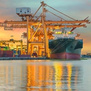 Large cargo ship docked at a port with towering orange cranes and stacked shipping containers, reflecting warm sunset colors on the water