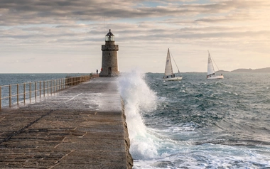 Stone pier with a lighthouse and waves crashing, two sailboats on the sea