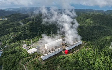 Aerial view of a geothermal power plant surrounded by green hills and steam.