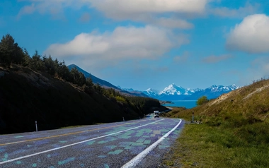 A rural highway winding through hills toward mountains and a lake under a blue sky