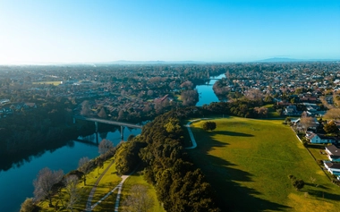 Aerial view of the Waikato River flowing through Hamilton, New Zealand, with riverside parkland, a bridge, and surrounding residential neighborhoods under a clear blue sky.